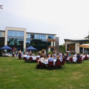 A group of families and students seated around circular tables on a grassy lawn in front of modern building