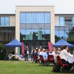 A group of families and students seated around circular tables on a grassy lawn in front of modern building